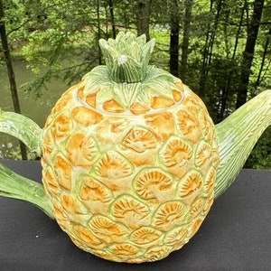 Può includere: Una teiera in ceramica a forma di ananas con foglie verdi e un design di ananas giallo e arancione.
