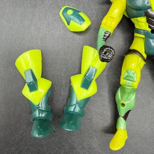 May include: A disassembled action figure with yellow and green armor. The figure's parts include two lower leg pieces, a head piece, and an arm. The figure's design features a combination of yellow and green colors.