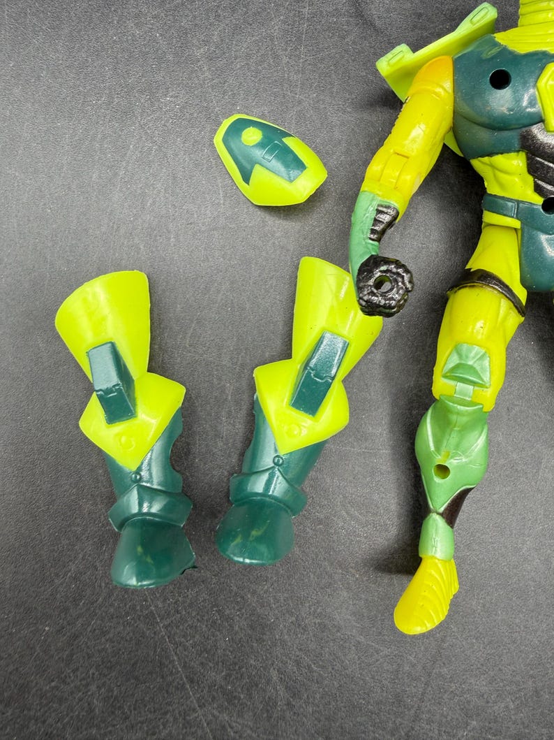 May include: A disassembled action figure with yellow and green armor. The figure's parts include two lower leg pieces, a head piece, and an arm. The figure's design features a combination of yellow and green colors.