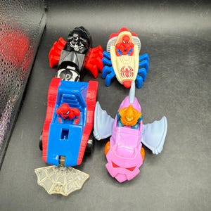 May include: Four colourful toy vehicles featuring Spider-Man characters. The toys include a black and red vehicle, a red and white vehicle, a blue and red vehicle, and a pink vehicle with blue wings. A clear plastic spiderweb is also included.
