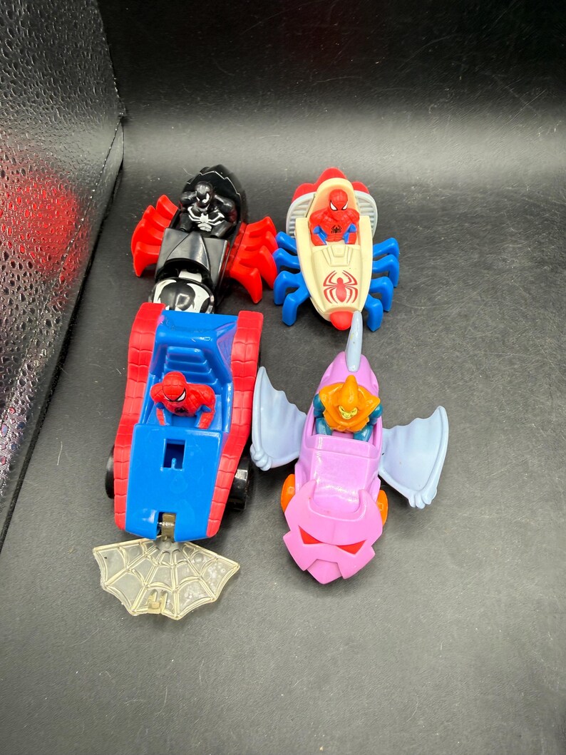 May include: Four colourful toy vehicles featuring Spider-Man characters. The toys include a black and red vehicle, a red and white vehicle, a blue and red vehicle, and a pink vehicle with blue wings. A clear plastic spiderweb is also included.