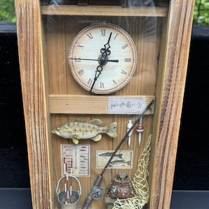 Puede incluir: Un reloj de pared con marco de madera con un tema de pesca. La esfera del reloj es blanca con números romanos negros y manecillas. El marco contiene una variedad de artículos relacionados con la pesca, incluyendo un pez, cañas de pescar, una red de pesca y un cubo.