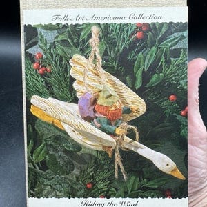 Puede incluir: Un adorno de madera que representa a un gnomo montando un ganso con las alas extendidas. El adorno está colgado de una cuerda y está rodeado de follaje verde. El texto "Folk Art Americana Collection" y "Riding the Wind" es visible en la caja.
