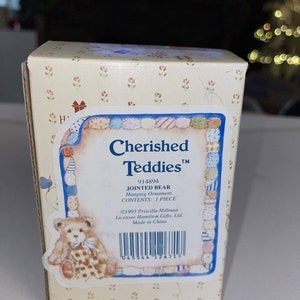 May include: A box for a Cherished Teddies™ 914894 jointed bear hanging ornament. The box is cream with a floral pattern and features a picture of a brown teddy bear wearing a plaid shirt and a bow. The text on the box reads "Cherished Teddies™ 914894 Jointed Bear Hanging Ornament Contents: 1 Piece ©1993 Priscilla Hillman Licensee Hamilton Gifts, Ltd. Made in China."