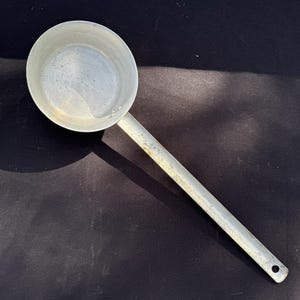 May include: A silver-coloured metal ladle with a round bowl and a long, straight handle. The handle has a small hole at the end for hanging. The ladle is likely used for cooking or serving food.