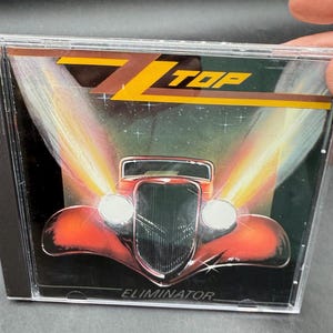 May include: A CD case featuring the album "Eliminator" by ZZ Top. The cover art showcases a red hot rod with bright headlights and the band's logo in yellow. The background has a starry, space-like design.