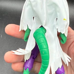 K&ouml;nnte beinhalten: Eine gr&uuml;ne und lila Actionfigur mit einem wei&szlig;en Umhang. Die Figur hat ein reptilienartiges Design mit einem langen gr&uuml;nen Schwanz, gr&uuml;nen Beinen und F&uuml;&szlig;en und einer lila Hose. Der wei&szlig;e Umhang hat einen zerfransten Rand.