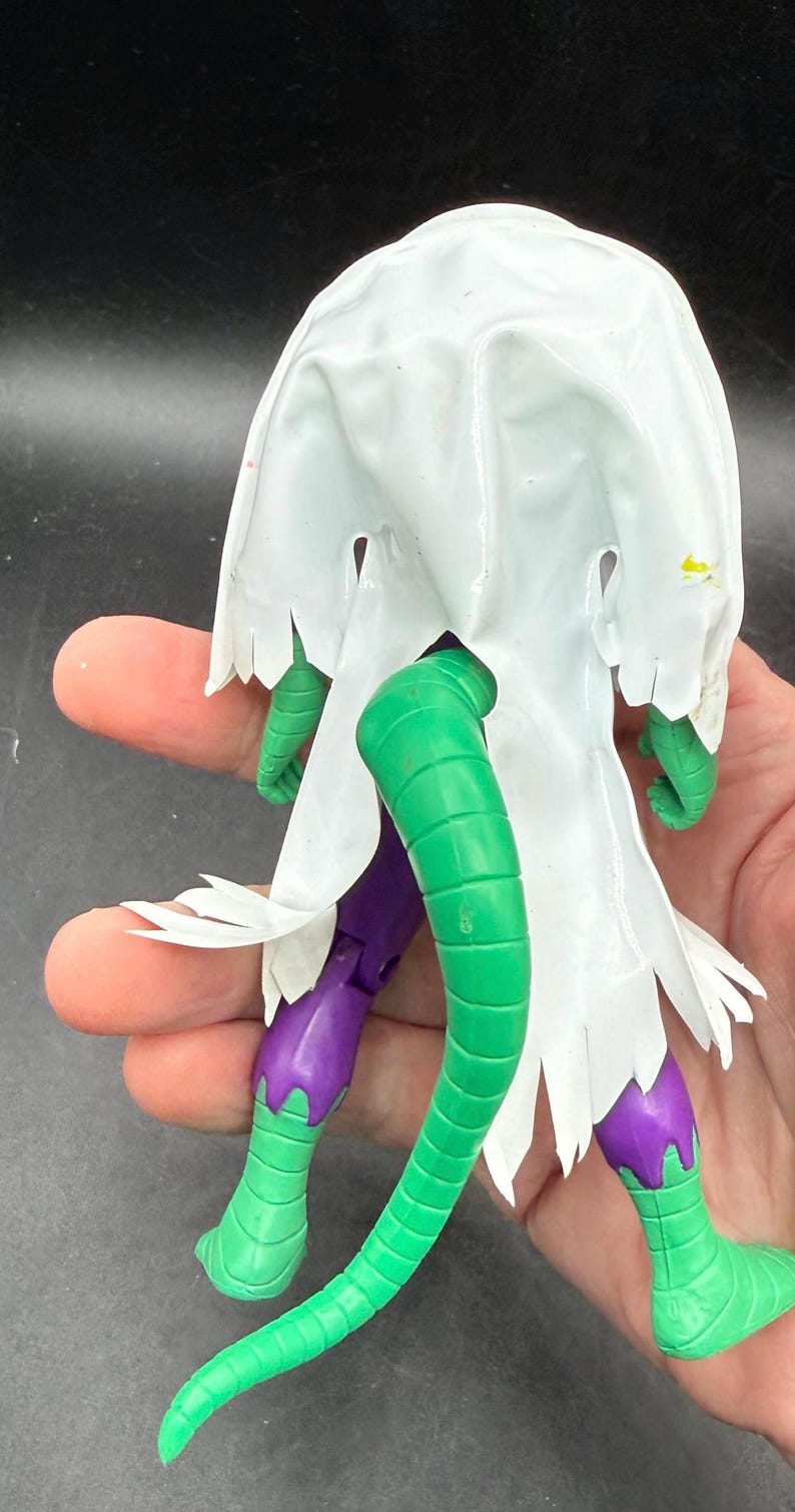 K&ouml;nnte beinhalten: Eine gr&uuml;ne und lila Actionfigur mit einem wei&szlig;en Umhang. Die Figur hat ein reptilienartiges Design mit einem langen gr&uuml;nen Schwanz, gr&uuml;nen Beinen und F&uuml;&szlig;en und einer lila Hose. Der wei&szlig;e Umhang hat einen zerfransten Rand.