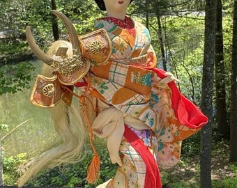 May include: A Japanese doll in a traditional kimono, featuring a detailed floral pattern in orange, red, and white. The doll has black hair and a decorative headpiece. It is standing on a black base, with a forest background.