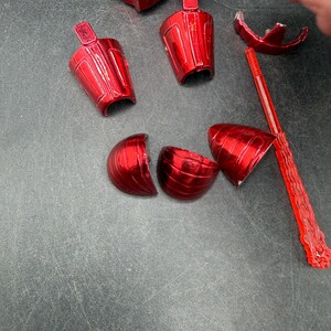 Puede incluir: Un conjunto de piezas de juguete rojas brillantes y desmontadas. Los componentes incluyen una estructura similar a una pistola, varias piezas en forma de copa y un elemento largo, delgado y segmentado. Las piezas est&aacute;n dispersas sobre una superficie gris oscuro.