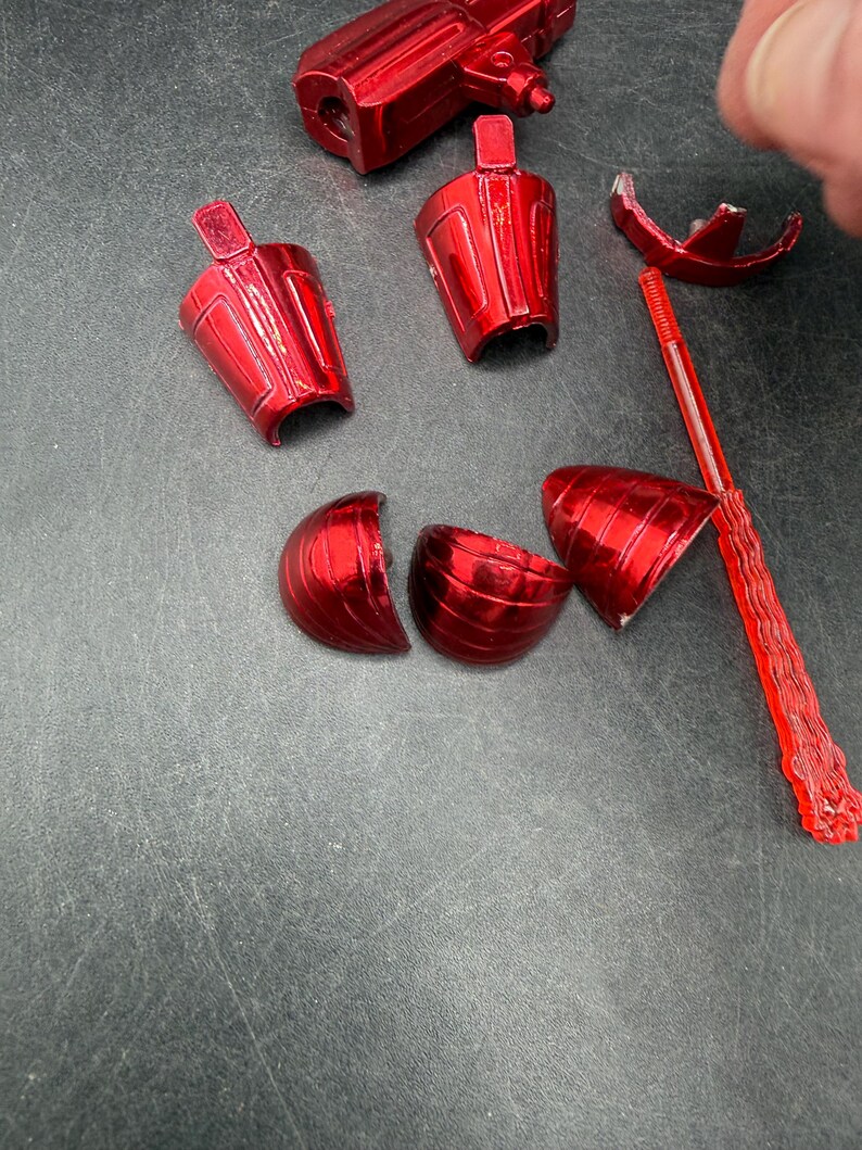 Puede incluir: Un conjunto de piezas de juguete rojas brillantes y desmontadas. Los componentes incluyen una estructura similar a una pistola, varias piezas en forma de copa y un elemento largo, delgado y segmentado. Las piezas est&aacute;n dispersas sobre una superficie gris oscuro.