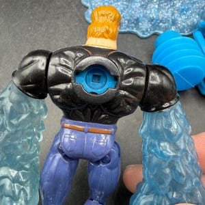 May include: A toy action figure with a black top, blue jeans, and blonde hair. The figure has blue water-like attachments on its arms. The figure is a superhero or villain character.
