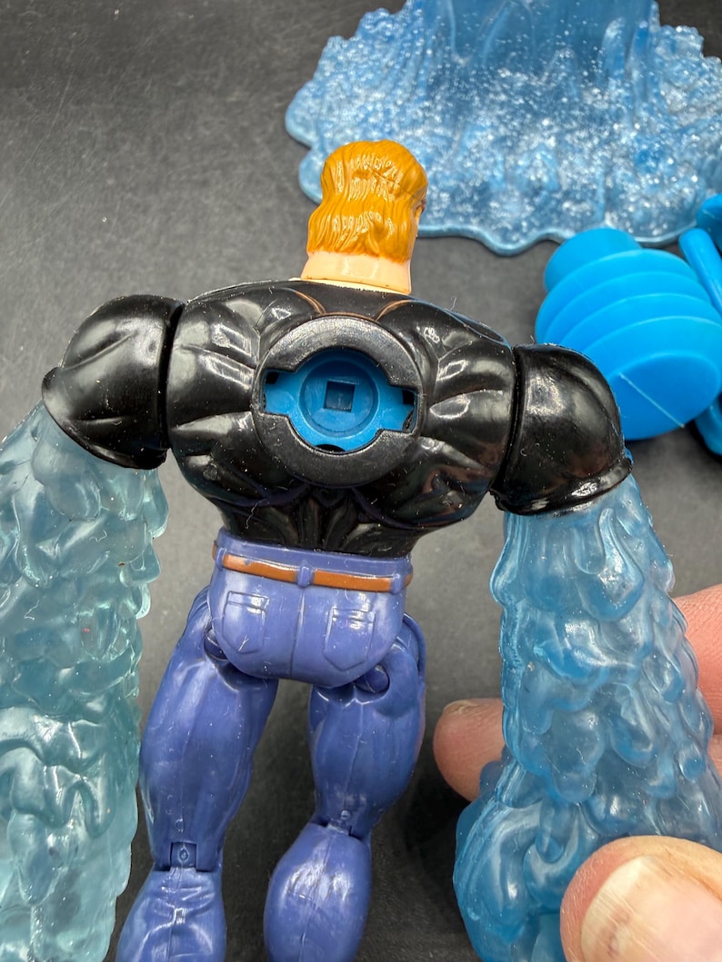 May include: A toy action figure with a black top, blue jeans, and blonde hair. The figure has blue water-like attachments on its arms. The figure is a superhero or villain character.