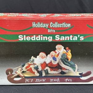May include: A red, green, and white box with a Christmas-themed image of three Santas on a sled. The box reads "Holiday Collection Gifts, Sledding Santa's, It's snow much fun".
