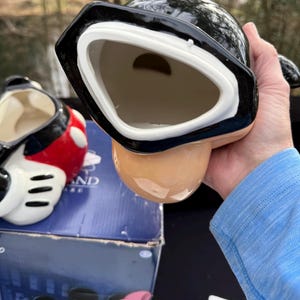 May include: A black and white ceramic mug with a unique design. The mug has a wide, open mouth and a rounded base. The interior is a light beige color. Another mug with red and black accents is visible in the background.