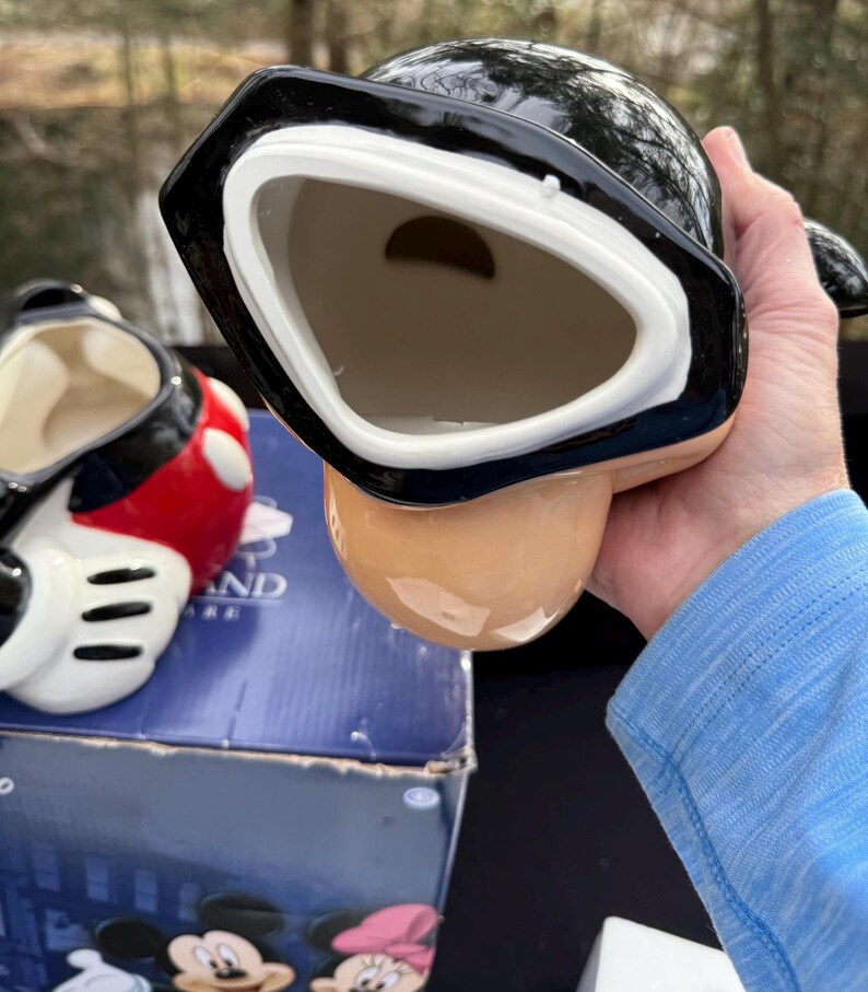 May include: A black and white ceramic mug with a unique design. The mug has a wide, open mouth and a rounded base. The interior is a light beige color. Another mug with red and black accents is visible in the background.