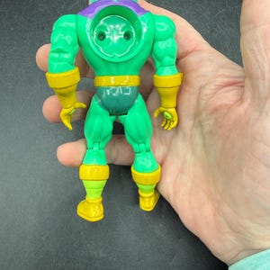 Op de afbeelding: Een groen en geel actiefiguur met een bolvormig hoofd, een paarse kraag en gele accenten op de armen, taille en benen. Het figuur wordt in een hand gehouden, wat het gedetailleerde ontwerp en de levendige kleuren benadrukt.