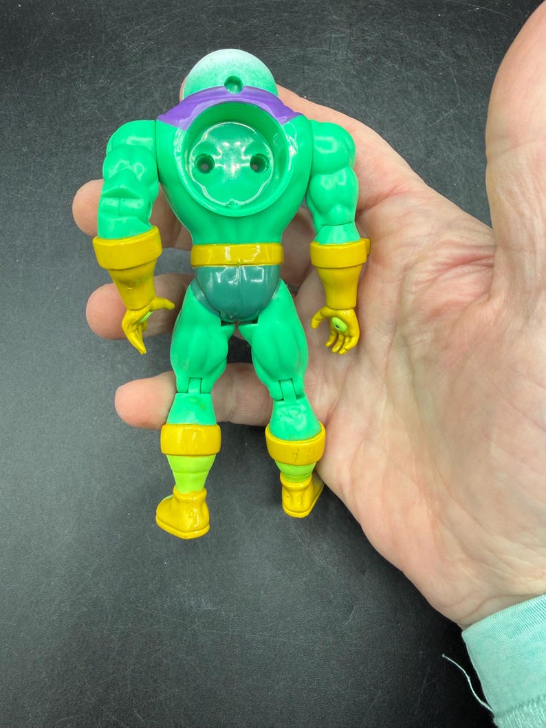 Op de afbeelding: Een groen en geel actiefiguur met een bolvormig hoofd, een paarse kraag en gele accenten op de armen, taille en benen. Het figuur wordt in een hand gehouden, wat het gedetailleerde ontwerp en de levendige kleuren benadrukt.