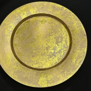 May include: A round, gold-colored charger plate with a textured surface and a wide rim. The plate has a metallic finish with yellow accents, creating a decorative and elegant look. The plate is designed for serving food.
