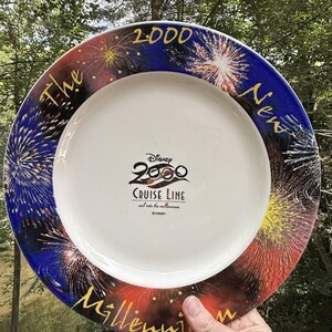 Puede incluir: Un plato redondo de cerámica blanca con un borde azul oscuro y rojo decorado con fuegos artificiales coloridos. El centro muestra el logotipo de Disney 2000 Cruise Line con el texto 'sail into the millennium'. El borde dice 'The New 2000 Millennium'.