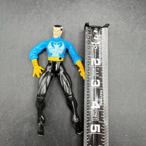 May include: A plastic action figure of a character in a blue and yellow costume with black pants. The figure is approximately 5 inches tall. The figure is standing next to a measuring tape.