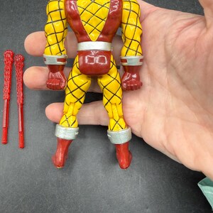 以下が含まれることがあります： 黄色と赤色のヴィンテージアクションフィギュア。黄色の部分にはひし形の模様があり、銀色のアクセントが付いています。2つの赤いプラスチック製のアクセサリーが含まれています。コレクション用の玩具です。