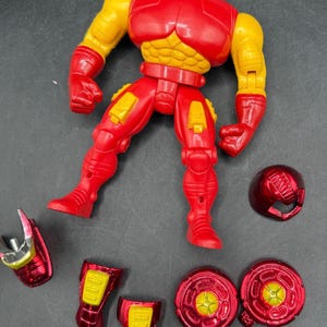 May include: A red and yellow action figure of a superhero, with detachable parts. The figure has a red helmet, chest, legs, and feet, with yellow arms and accents. Additional red and yellow pieces are scattered around the figure.