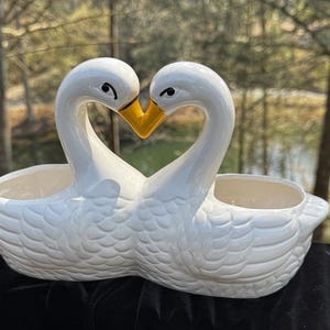 May include: A white ceramic double planter shaped like two swans facing each other with their necks forming a heart. The swans have glossy white bodies with molded feather details and bright yellow beaks. The piece features two open compartments.