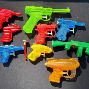 May include: A collection of colorful plastic toy water guns in red, green, blue, and yellow. The water guns are in various shapes and sizes, with a retro aesthetic. The toys are arranged on a dark surface.