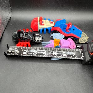 May include: A selection of colourful toy vehicles featuring Spider-Man characters. The toys include a red and blue car, a black spider-like vehicle, and a pink and purple vehicle. A measuring tape is placed next to the toys.