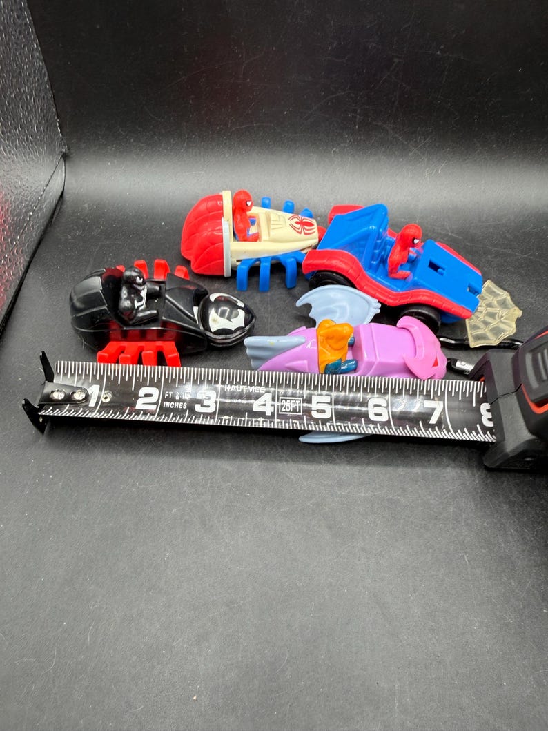 May include: A selection of colourful toy vehicles featuring Spider-Man characters. The toys include a red and blue car, a black spider-like vehicle, and a pink and purple vehicle. A measuring tape is placed next to the toys.