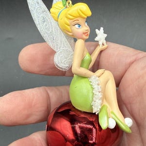 May include: A Tinker Bell figurine with blonde hair in a bun, wearing a green dress with white trim, and translucent glittery wings. The character sits atop a large, shiny red jingle bell and holds a small white snowflake in one hand.