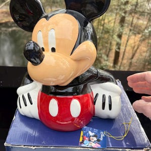 May include: A ceramic Mickey Mouse figurine with black ears, a tan face, and a red and white body. The figurine is on a blue Disney box. The box has the Disney logo and a tag with Mickey and Minnie Mouse.