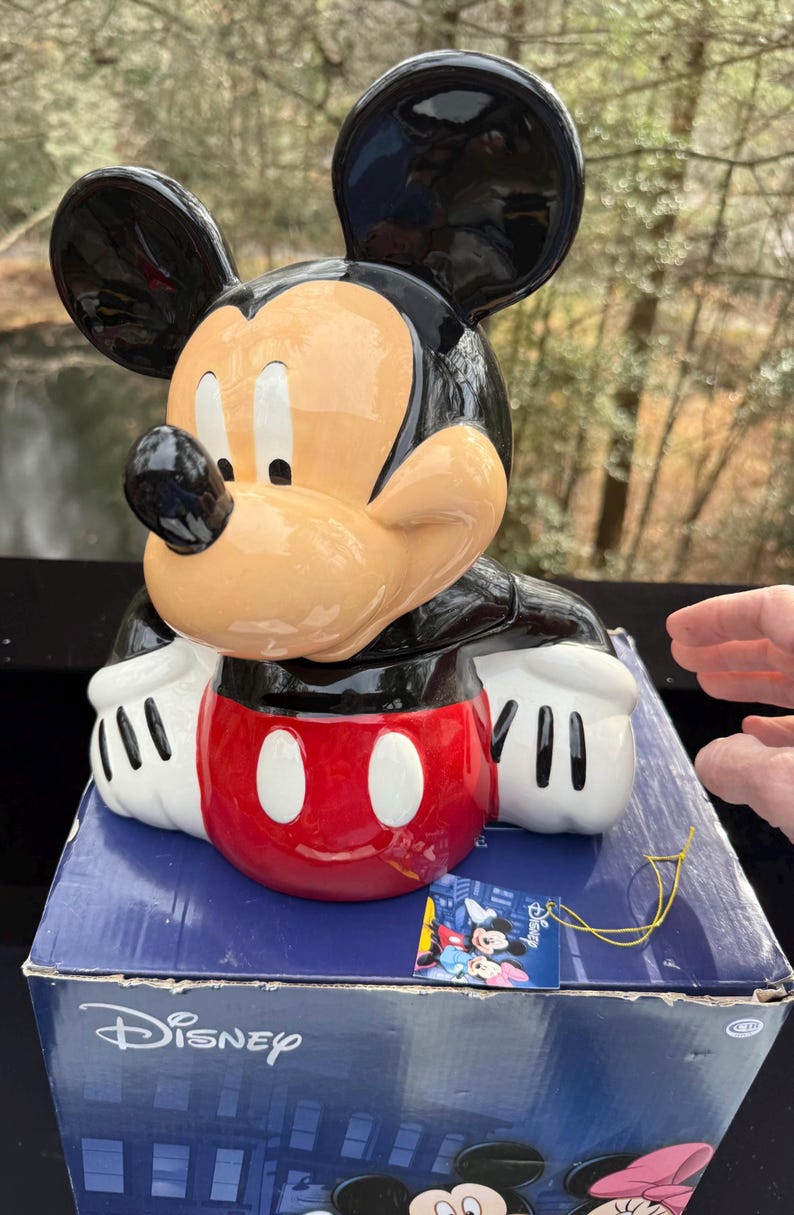 May include: A ceramic Mickey Mouse figurine with black ears, a tan face, and a red and white body. The figurine is on a blue Disney box. The box has the Disney logo and a tag with Mickey and Minnie Mouse.