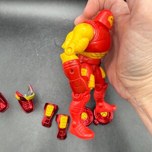 May include: A red and yellow Iron Man action figure with detachable parts. The figure has a red helmet, torso, and legs, with yellow arms and accents. Several red and yellow pieces are detached, including boots and a faceplate.