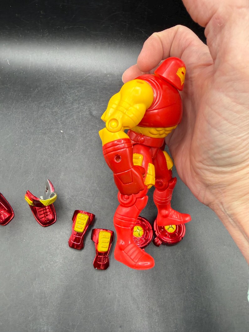 May include: A red and yellow Iron Man action figure with detachable parts. The figure has a red helmet, torso, and legs, with yellow arms and accents. Several red and yellow pieces are detached, including boots and a faceplate.