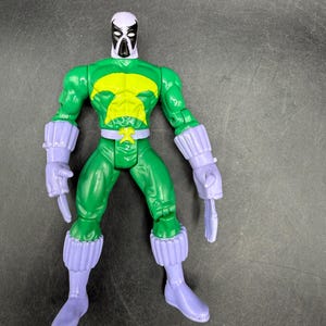 May include: A green action figure with a yellow chest emblem, purple gloves, boots, and a mask with a white head and black face. The figure is posed with its arms at its sides and two purple blades in its hands.