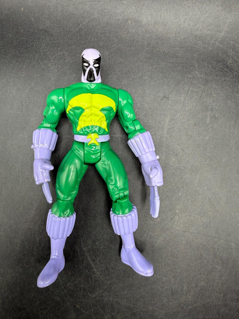 May include: A green action figure with a yellow chest emblem, purple gloves, boots, and a mask with a white head and black face. The figure is posed with its arms at its sides and two purple blades in its hands.