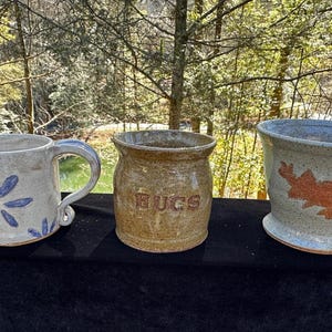 May include: Three handmade ceramic pieces: a mug with blue flower designs, a small jar with the word "HUGS", and a flared bowl with an orange floral pattern. The pottery is in shades of beige and blue.
