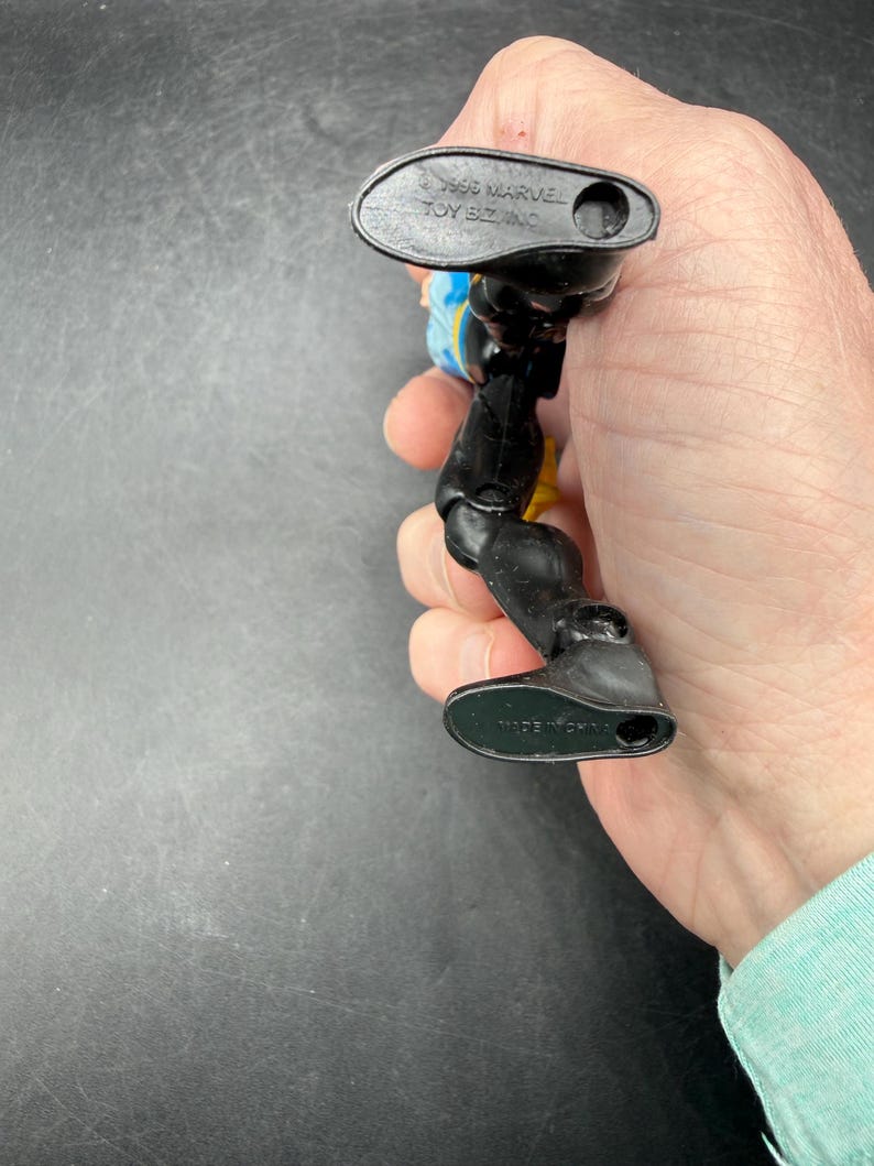 May include: A black and yellow action figure with blue accents. The figure is held in a hand, with the feet of the figure visible. The figure is marked with "&copy; 1996 MARVEL TOY BIZ, INC." and "MADE IN CHINA."