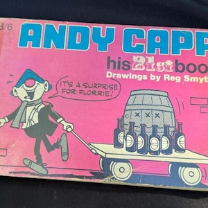 May include: A vintage comic book titled "Andy Capp his 21st book" with drawings by Reg Smythe. The cover features a cartoon character pulling a cart of beer bottles and a barrel. The background is pink with blue text.