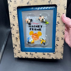May include: A blue magnet frame from The Disney Store, with four character magnets. The frame is in a cardboard box with a Mickey Mouse pattern. The frame text reads "MAGNET FRAME" and "HOLDS: photos, notes, cards."