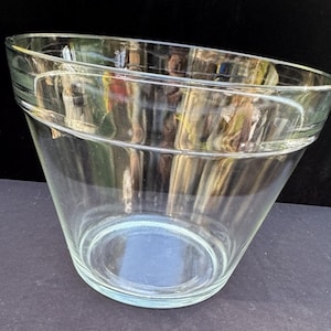 May include: A clear glass bowl with a wide, flared rim and a tapered base. The vessel features a smooth, transparent surface with a distinct horizontal ridge encircling the upper section, providing a simple, functional design for various uses.