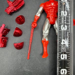 Puede incluir: Figura de acci&oacute;n de Iron Man roja y plateada, de aproximadamente 15 cm de altura, con piezas de armadura desmontables. La figura tiene torso y extremidades rojas, piernas y brazos plateados, y un arma roja. La figura est&aacute; en posici&oacute;n vertical.