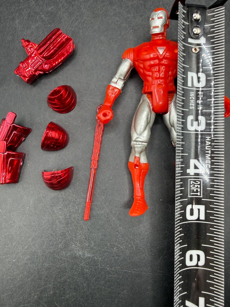 Puede incluir: Figura de acci&oacute;n de Iron Man roja y plateada, de aproximadamente 15 cm de altura, con piezas de armadura desmontables. La figura tiene torso y extremidades rojas, piernas y brazos plateados, y un arma roja. La figura est&aacute; en posici&oacute;n vertical.