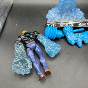 May include: A disassembled action figure with blue water-like arms and a matching head. The figure has purple pants, brown boots, and a black torso. Additional blue water-like accessories are also present, along with a ruler.