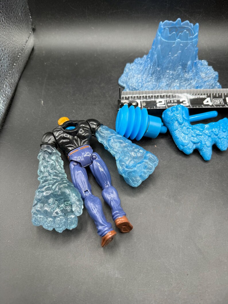 May include: A disassembled action figure with blue water-like arms and a matching head. The figure has purple pants, brown boots, and a black torso. Additional blue water-like accessories are also present, along with a ruler.