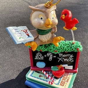 May include: A Christmas ornament featuring a brown owl wearing a green scarf, holding a book that says "Santa Claus is coming to town". The owl is perched on a stack of books with a chalkboard that says "Merry Xmas" and a red bird flying above.