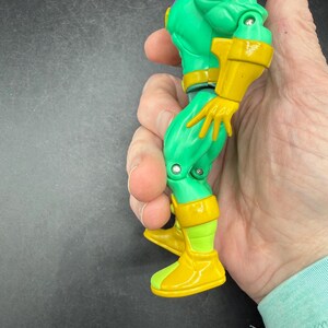 Op de afbeelding: Een groen en geel actiefiguur met een helm en gele laarzen. Het figuur heeft een gespierde bouw en wordt in een hand gehouden. Het ontwerp van het figuur bevat gele accenten op de armen en benen.