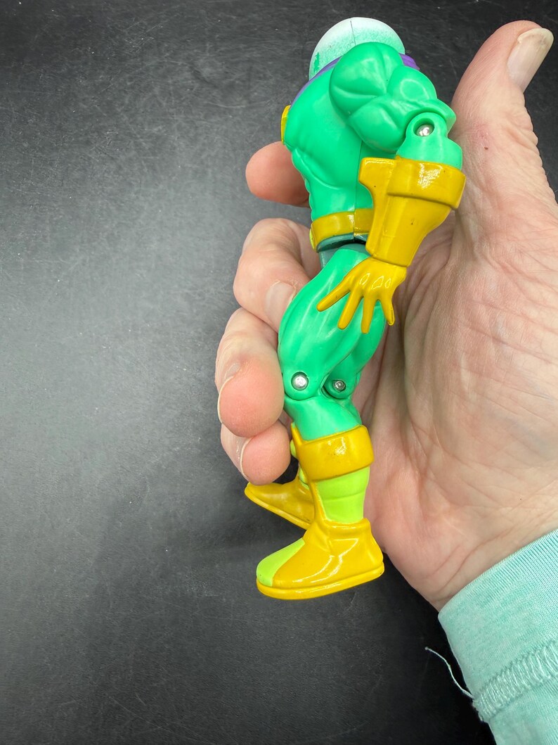 Op de afbeelding: Een groen en geel actiefiguur met een helm en gele laarzen. Het figuur heeft een gespierde bouw en wordt in een hand gehouden. Het ontwerp van het figuur bevat gele accenten op de armen en benen.
